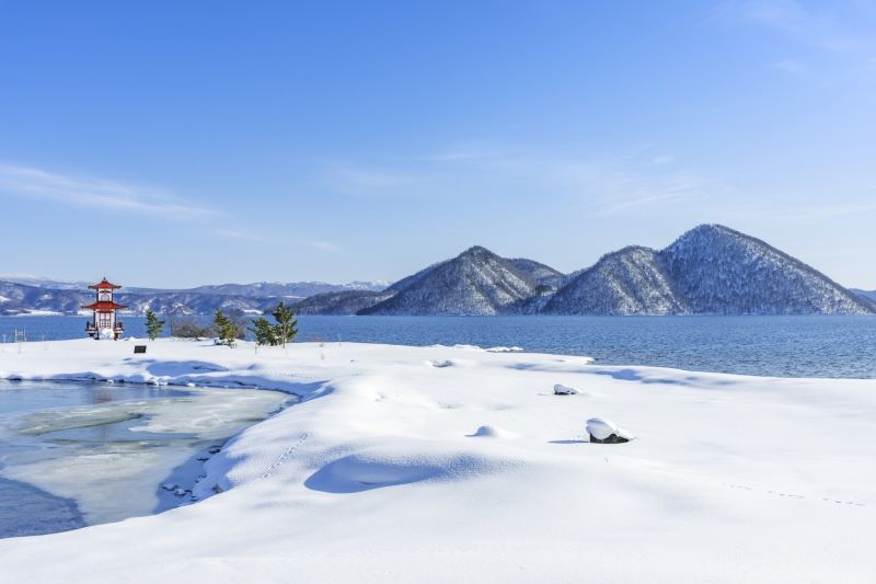 A winter landscape of Lake Toya in Hokkaido, with snow-covered shores, a small red and white lighthouse or observation tower, and snow-capped mountains rising from the blue water under a clear sky