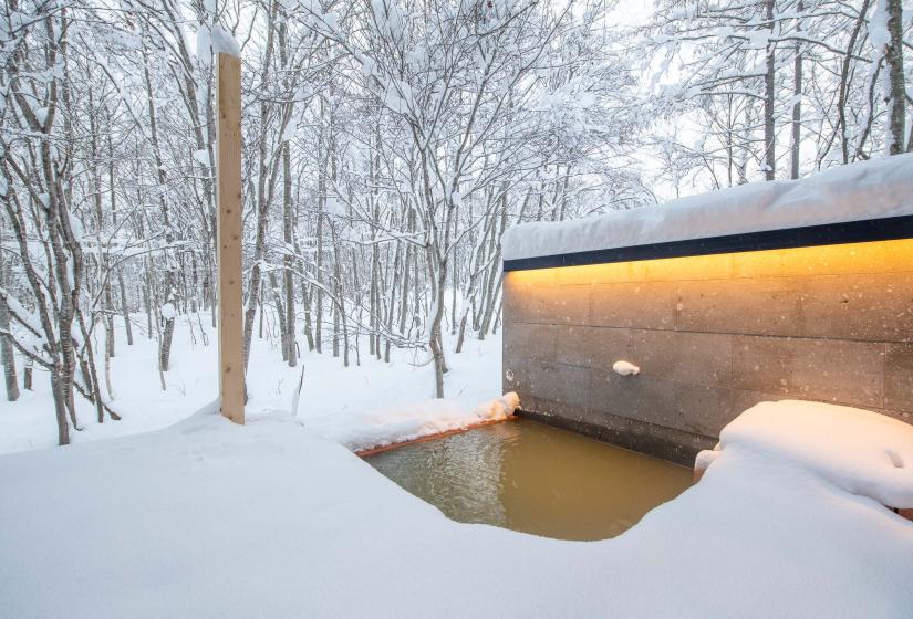 Private outdoor onsen at Moraine Chalet featuring a modern rectangular bath with minimalist concrete walls and warm lighting, surrounded by deep snow and frost-covered birch trees in winter