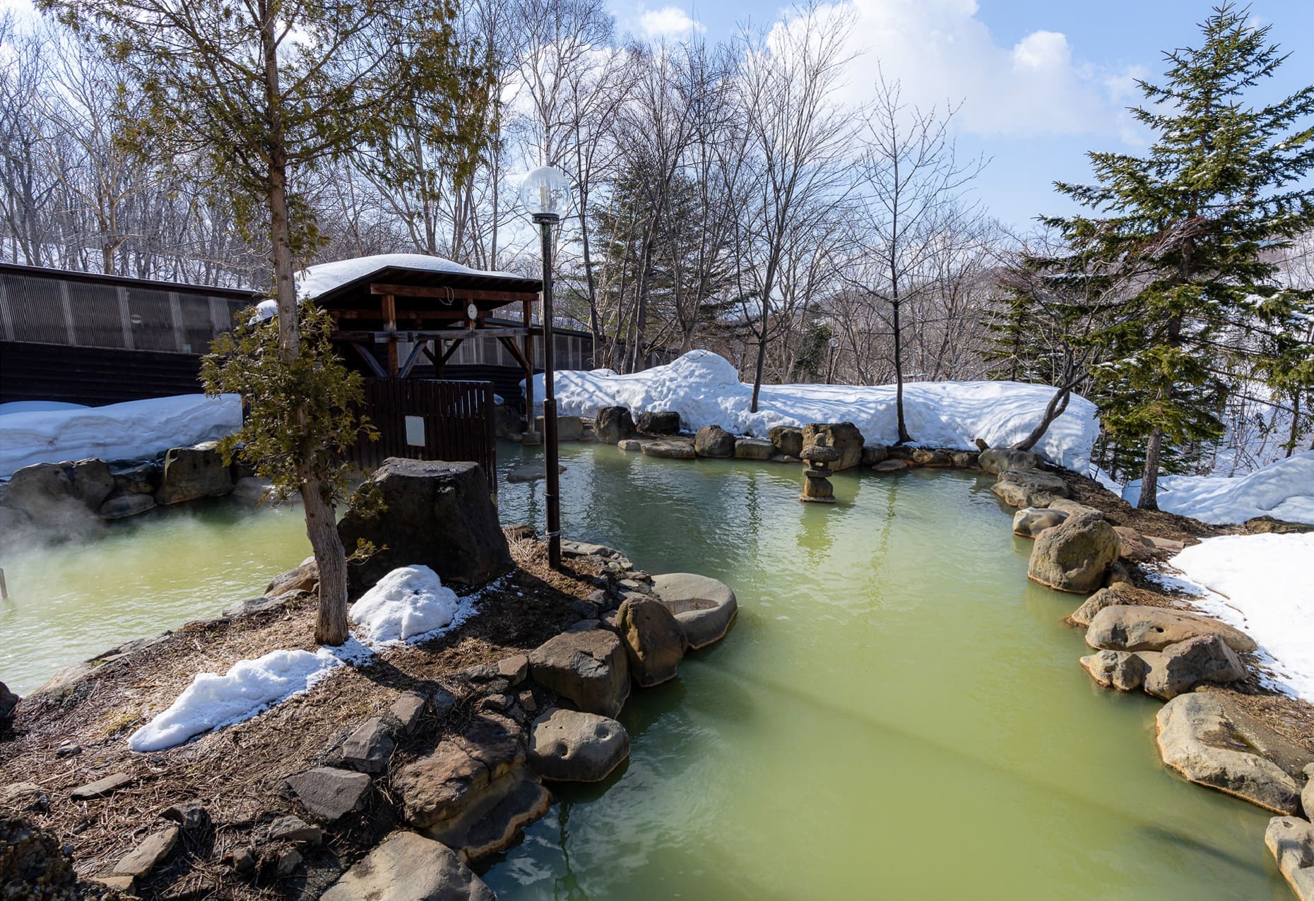 Outdoor mixed onsen at Niseko Grand Hotel featuring sulfur-tinted green waters surrounded by natural rocks and snow banks, with a wooden covered rest area and evergreen trees in a winter landscape