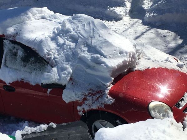 A red car partially buried under a large pile of fallen snow, likely from a roof or overhang, causing significant coverage of the vehicle