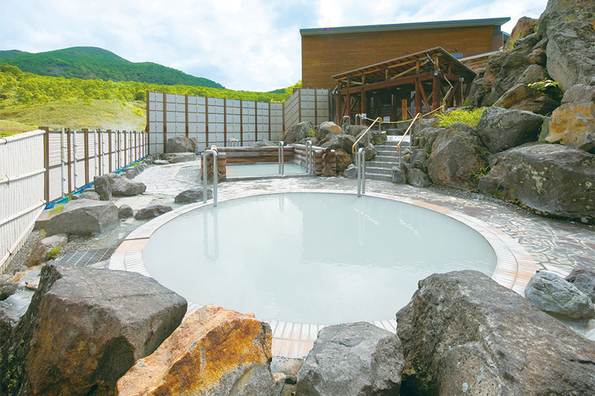 Outdoor onsen bath at Goshiki Onsen featuring a circular milky-white mineral pool surrounded by large volcanic rocks, wooden decking, and handrails, set against green summer mountains and traditional Japanese architecture