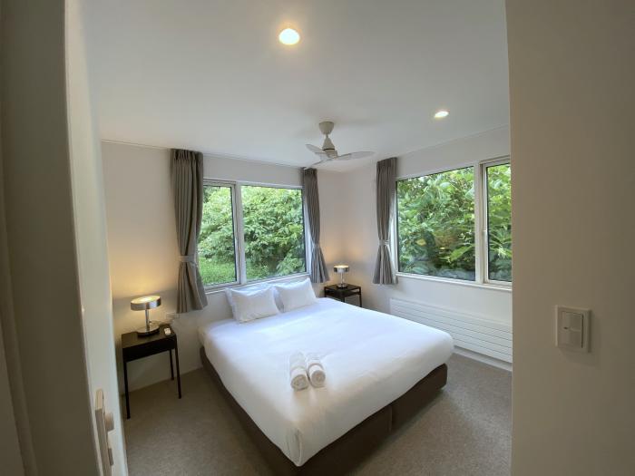 A bedroom with open curtains and 2 large windows with leafy surrounds
