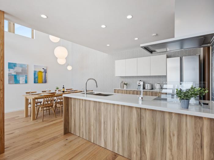Modern open-plan kitchen and dining space with light wood cabinetry, white countertops, and stainless steel appliances. Natural light streams in through high windows, illuminating the wooden dining table and chairs in the background