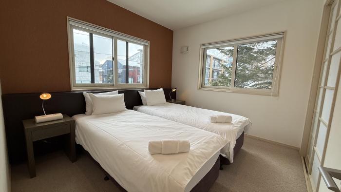 Stylish twin bedroom at Yutaka Townhouses with two comfortable beds dressed in white linens, bedside tables with reading lamps, large windows overlooking snowy Niseko forest, and warm wood accent wall creating a cozy mountain retreat atmosphere.