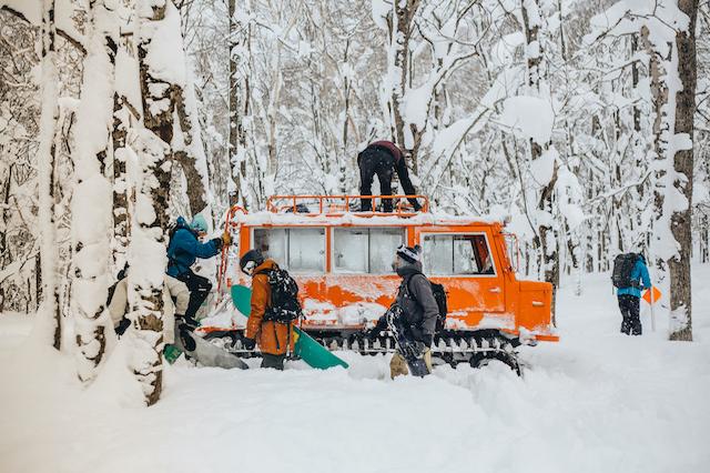 Skiers and snowboarders alight from an orange snow cat in snowy trees