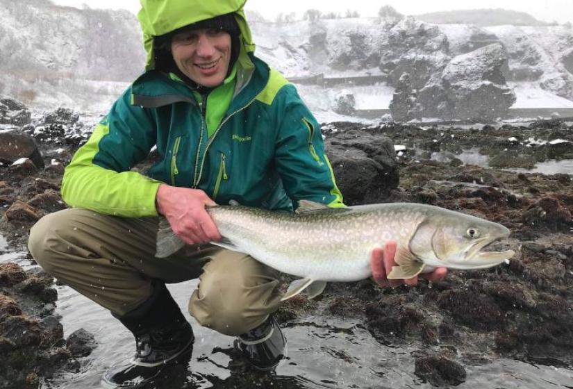 A man holds a spotted char with snow in the back ground