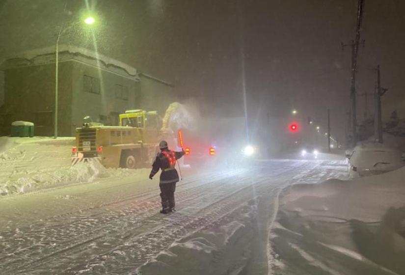 Man directs traffic with a glowing stick while a snow removal machine operates