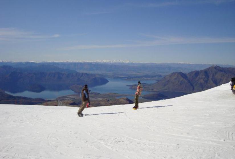 Two snowboarders ride down a slope with a beautiful lake back drop