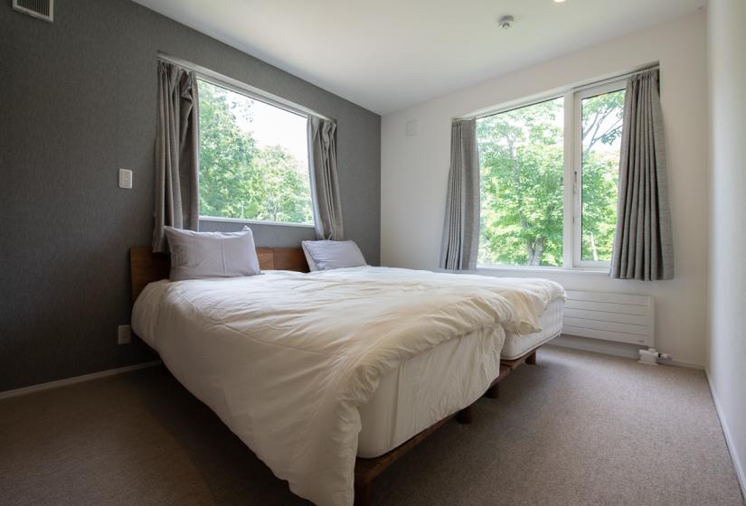 A made up double bed  in a brown carpeted room with windows behind.