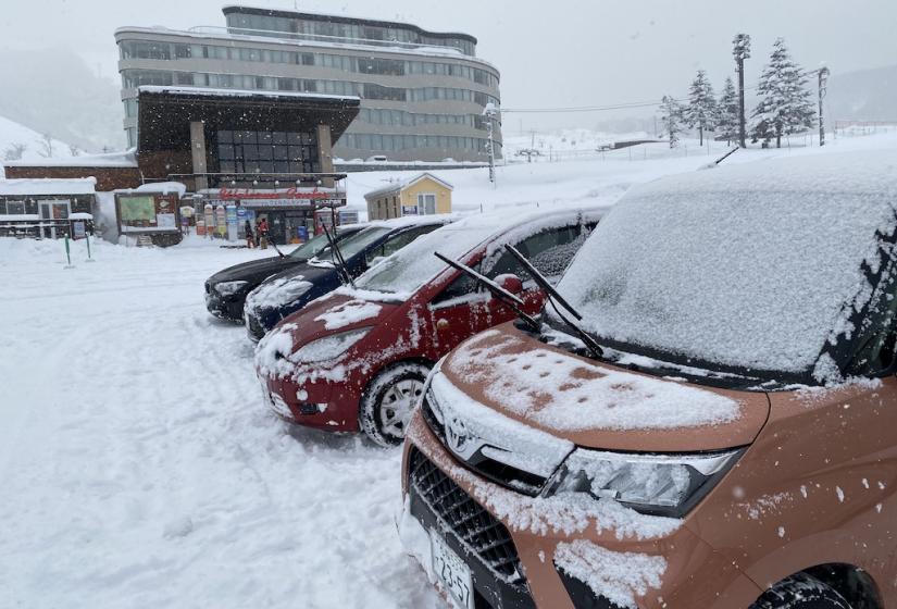 A row of cars with their wipers up and the Hirafu Welcome Center in the back ground