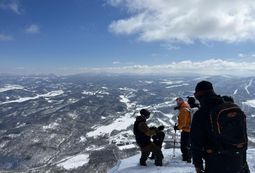 Imagine being dropped off on the top of a peak with a group of your best friends on the sunniest of days with fields of the deepest lightest powder you can find below. A group of snowboarders on top of a peak with a view to the valley below.