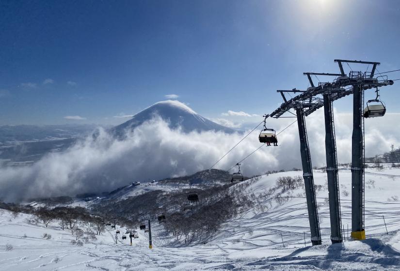 We can't wait to welcome you back for more incredible days like these. Blue skies above a chairlift tower with Mount Yotei in the back ground.