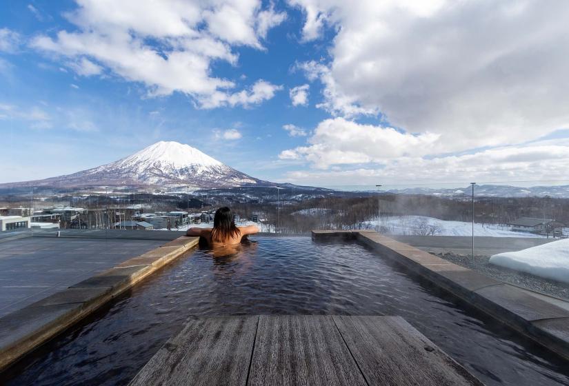 A person sitting in an pool with Mount Yotei in the background - Aya Niseko | FintechZoom
