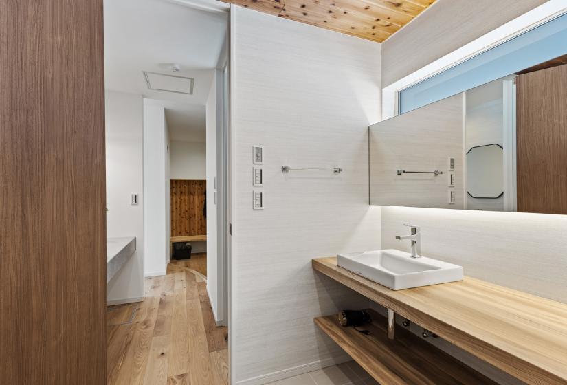 Modern bathroom with wooden floating vanity, wide mirror, and natural wood ceiling feature. Long corridor view shows access to additional bathroom spaces, with bright white walls contrasting warm wood elements