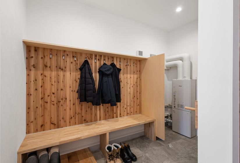 Ski room entry area with natural wood wall paneling featuring coat hooks, a built-in bench, and shoe storage space below. The minimalist design includes concrete flooring and bright white walls, with winter jackets displayed on hooks