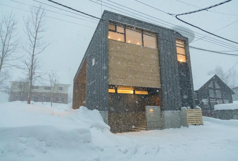 Dramatic exterior view of modern charcoal-colored chalet during heavy snowfall, with warm light glowing from windows. The contemporary angular architecture stands out against deep snow banks, creating a cozy winter atmosphere in Niseko's snowscape.