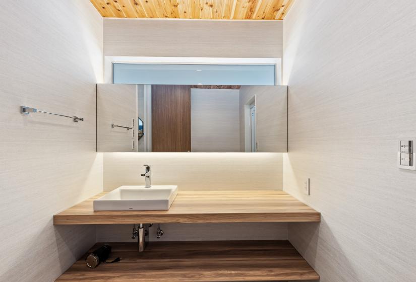 Modern bathroom vanity featuring a floating wooden counter, wall-mounted sink, and full-width mirror with overhead natural lighting through a skylight. The minimalist design combines white walls with natural wood tones