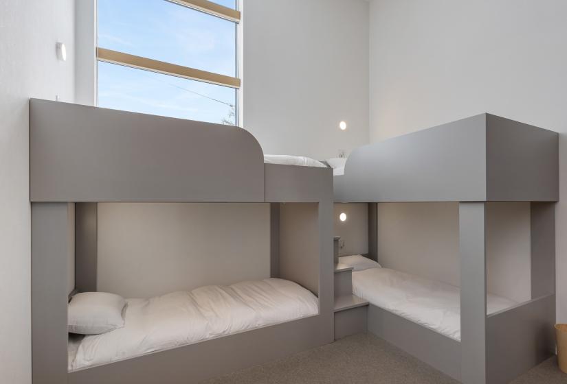 Close-up view of custom-designed bunk beds in grey finish with white bedding. The modern, minimalist bunks feature clean lines and a contemporary design, ideal for families or groups. Natural light enters through a tall window