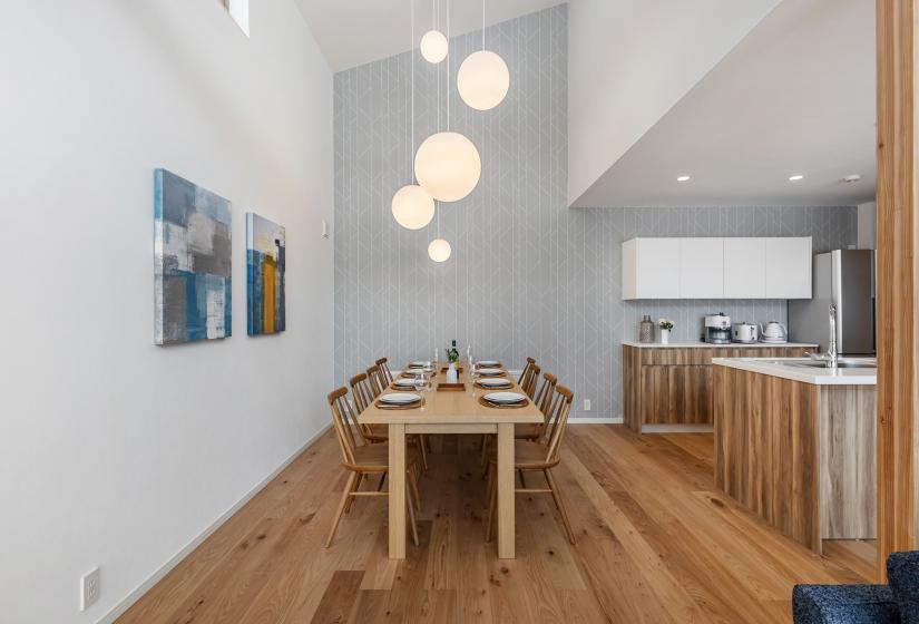 Elegant dining space with natural wood table seating eight guests, suspended globe pendant lights, and modern artwork on white walls. Hardwood flooring extends to the open kitchen featuring light wood cabinetry