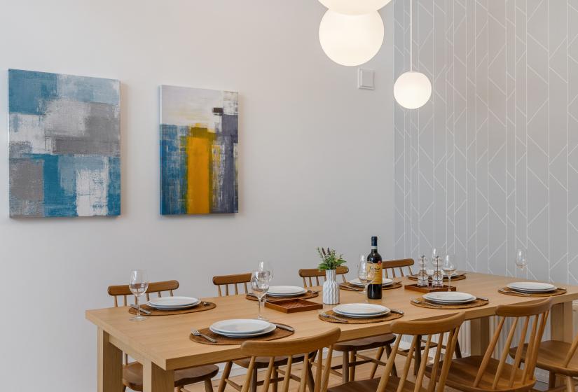 Dining table set for a meal with wooden chairs, contemporary globe light fixtures overhead, and colorful abstract artwork adorning the white walls. The wooden table offers comfortable seating for group dining.