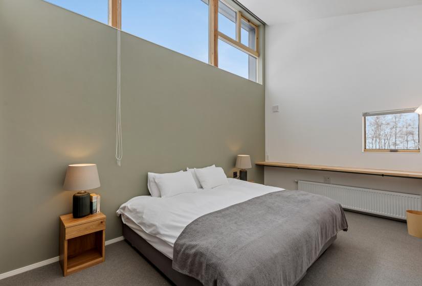 Master bedroom with double-size bed dressed in white linens and grey throw, featuring a sage green accent wall, wooden floating shelf, and high windows for natural light. Modern bedside table with lamp completes the contemporary design