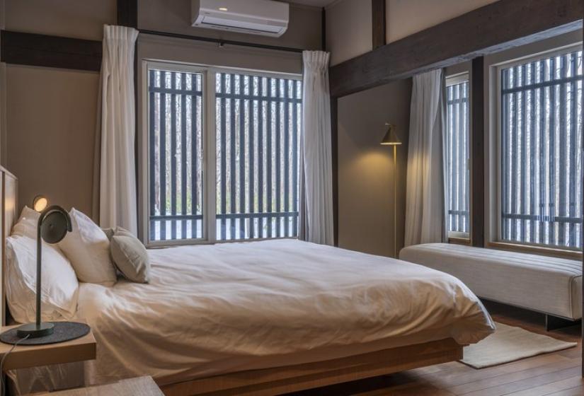 Serene bedroom at Nagare House featuring a comfortable king-sized bed with crisp white bedding, wooden flooring, and large traditional Japanese windows with vertical slats that filter natural light. The room combines authentic Japanese architectural elements with modern comfort, perfect for a luxury ski vacation in Niseko, Japan.