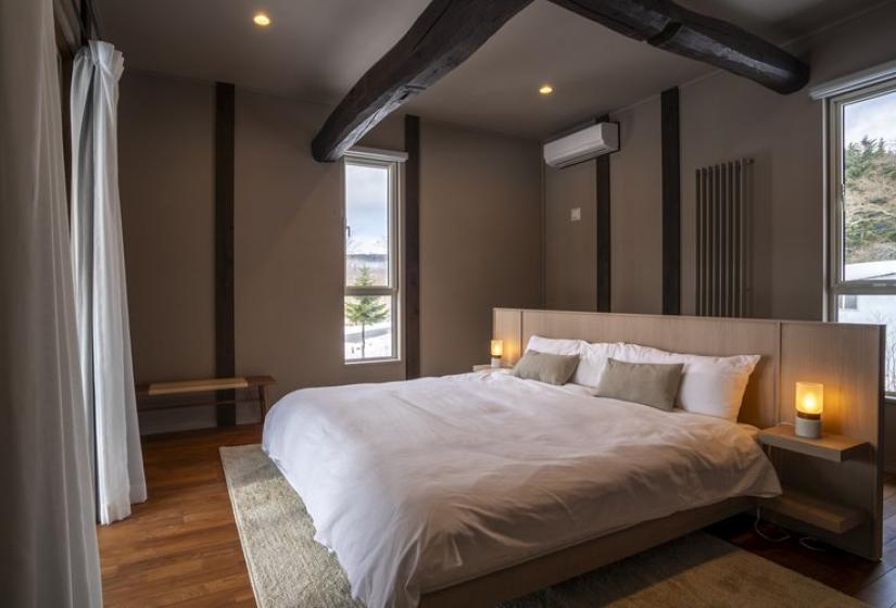 A stylish modern bedroom with a comfortable king-sized bed featuring white linens, wooden flooring, exposed wooden ceiling beams, and large windows letting in natural light. The room has a warm, minimalist design with neutral tones and includes bedside lamps for ambient lighting