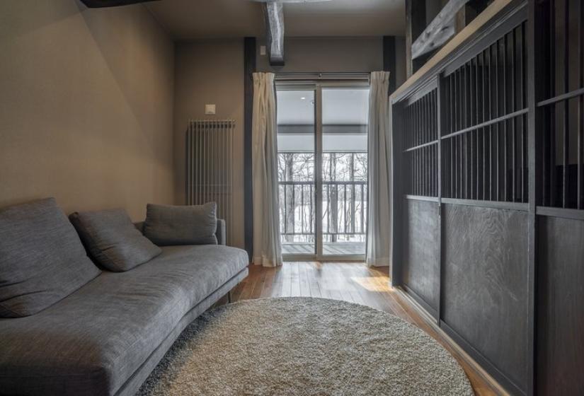 A cozy living area featuring a gray sofa with cushions positioned against the wall, a round area rug on wooden flooring, and sliding glass doors that open to a balcony bringing in natural light. The room has traditional Japanese architectural elements with wooden built-in cabinetry and neutral-toned walls, creating a serene, minimalist atmosphere.