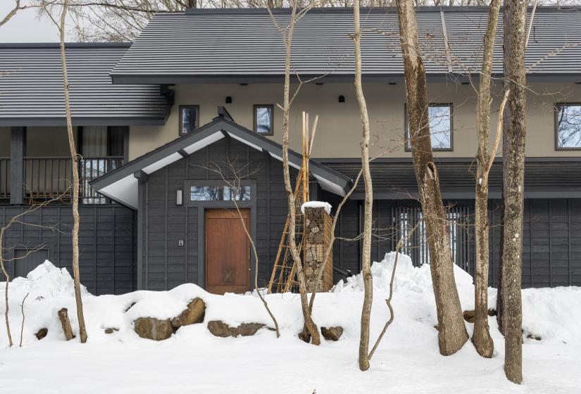Exterior view of Nagare House featuring its charming traditional Japanese architecture with dark wooden façade and sloped roof, surrounded by snow-covered ground and bare winter trees. This beautifully restored 100-year-old kominka farmhouse sits nestled in a serene woodland setting, offering an authentic Japanese experience just minutes from Niseko's ski resorts.