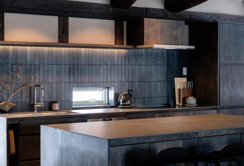 Elegant kitchen at Nagare House featuring a sleek dark island countertop, contemporary blue-gray tiled backsplash, minimalist wooden cabinetry, and traditional exposed wooden beam ceiling. The beautifully designed kitchen combines modern functionality with traditional Japanese architectural elements, perfect for self-catering during your luxury ski vacation in Niseko.