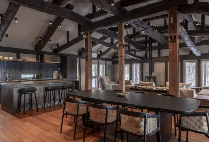 Sophisticated open kitchen and bar area at Nagare House featuring a central island with bar seating, wooden dining table with modern black chairs, and striking exposed wooden beam architecture. The restored 100-year-old kominka farmhouse blends traditional Japanese structural elements with contemporary kitchen design, creating an elegant space for social dining during your luxury ski holiday in Niseko.
