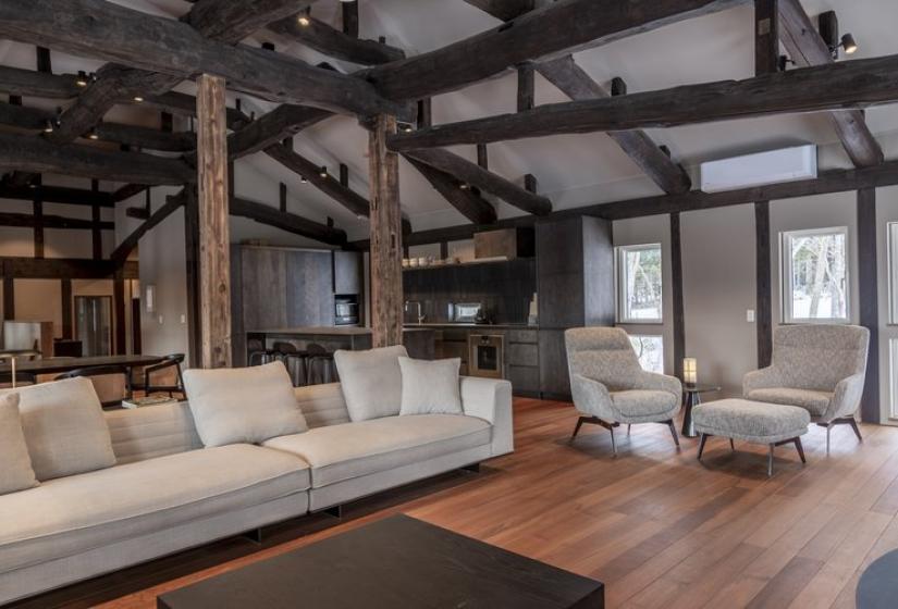 Stunning open-concept living room at Nagare House featuring a comfortable white sectional sofa, wooden coffee table, plush armchair, and magnificent exposed wooden beam architecture. The beautifully restored 100-year-old kominka farmhouse combines traditional Japanese structural elements with modern comfort, creating an inviting space for relaxing after skiing in Niseko, with natural light streaming through multiple windows.
