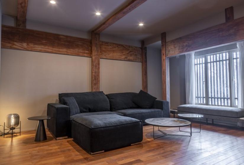 Modern living room at Nagare House featuring a spacious dark gray sectional sofa, glass coffee table, warm wooden floors, exposed wooden beams, and traditional Japanese latticed windows bringing in natural light. The perfect relaxation space for luxury ski accommodation in Niseko, blending contemporary comfort with authentic Japanese architectural elements.