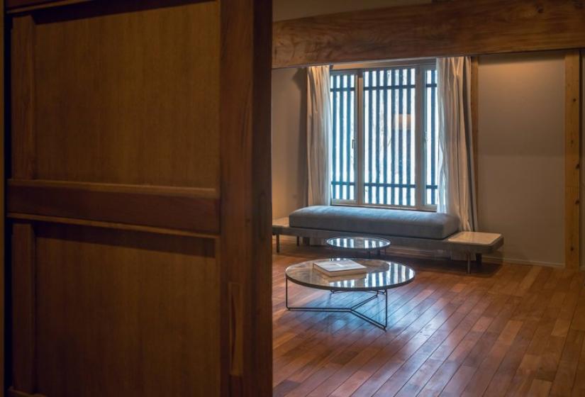 Glimpse through wooden sliding doors into a tranquil tea room at Nagare House featuring a traditional Japanese round table, polished wooden floors, and authentic Japanese-style latticed windows that filter soft natural light, showcasing the 100-year-old kominka farmhouse heritage of this luxury Niseko ski accommodation.