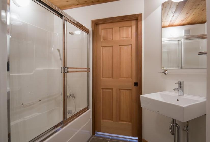 A clean bathroom with wooden ceiling, glass shower enclosure, white sink, and wooden door. The space features neutral tiled flooring, white walls, and warm overhead lighting creating a functional and comfortable bathroom area.