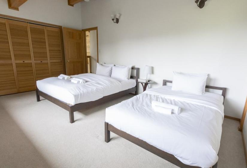 A clean, minimalist bedroom with two beds featuring crisp white linens and dark wooden frames. The room has light-colored carpeting, white walls, wooden ceiling beams, and built-in storage, creating a simple and peaceful sleeping space.