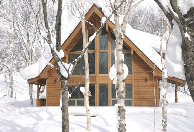 Rustic wooden A-frame cabin in snowy Niseko winter landscape, featuring large glass windows and traditional Japanese alpine architecture surrounded by bare birch trees