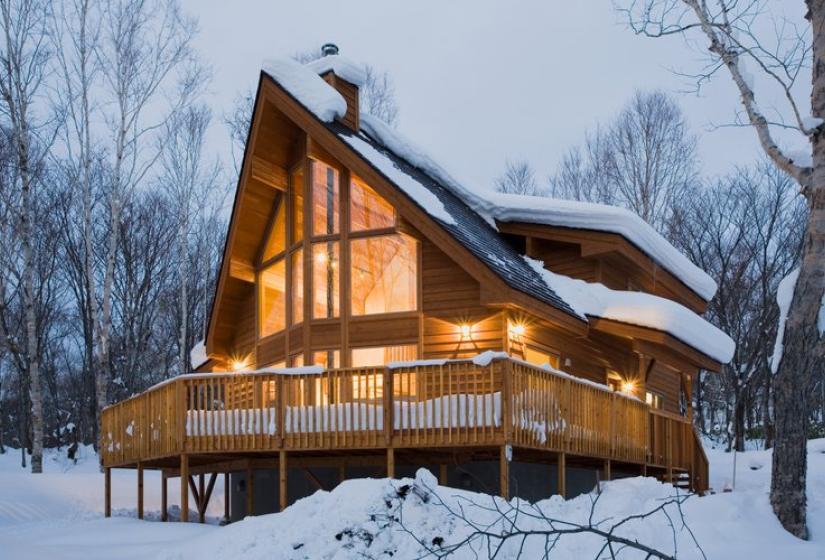 A modern wooden A-frame cabin with large glass windows illuminated from within, surrounded by snow and bare trees in Annupuri, Niseko. The cabin features a wraparound deck built on stilts and a distinctive triangular facade glowing warmly against the twilight winter landscape.