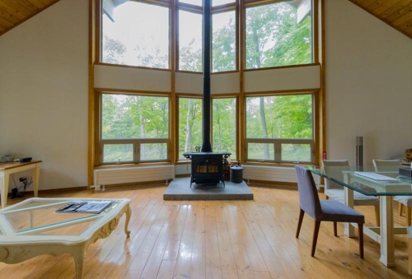 A bright, open-plan living space with large floor-to-ceiling windows showcasing forest views. The room features wooden flooring, a central wood-burning stove, minimalist furniture, and a dining table, all beneath a wooden cathedral ceiling with natural light flooding the interior.