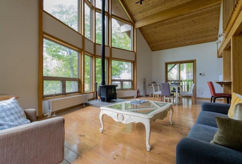 A spacious living room with high ceilings, large windows, and wooden beams. The room features a comfortable seating area with a sofa, armchair, and coffee table on wooden flooring. Natural light streams in through multiple windows showcasing forest views outside.
