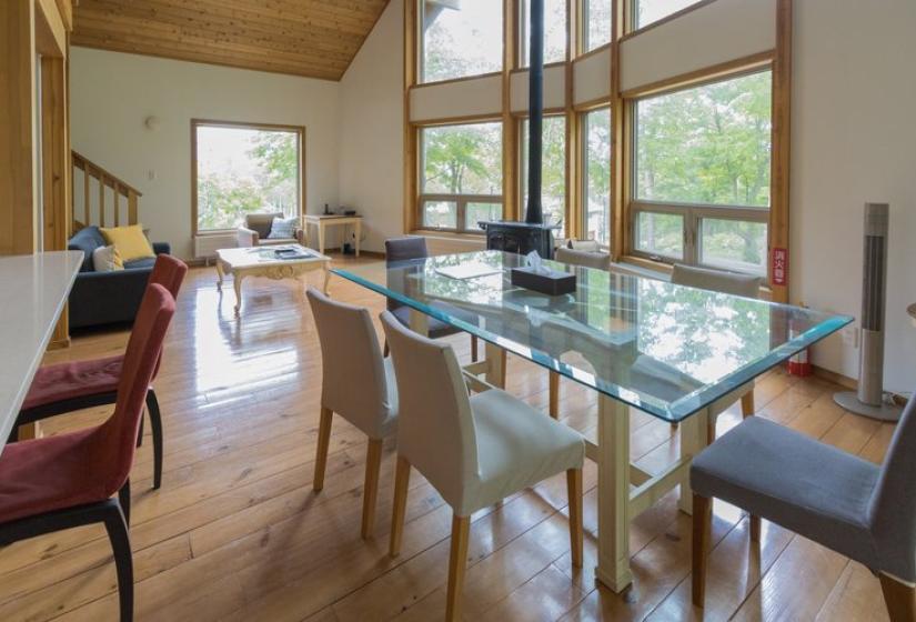 A bright dining area with a glass dining table surrounded by stylish chairs of different designs. The space features wooden flooring, large windows with forest views, wooden ceiling, and an open-concept layout connecting to the kitchen and living areas.