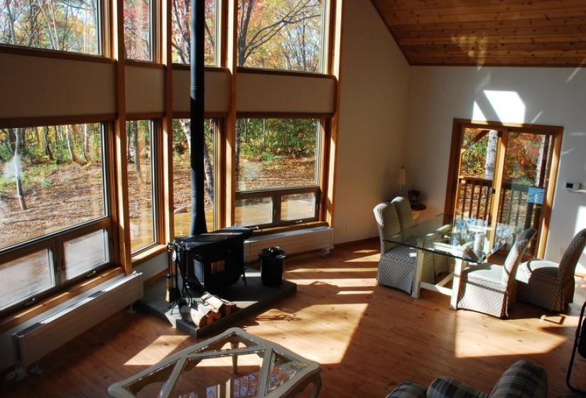 A bright corner living area with floor-to-ceiling windows showcasing scenic forest views. The space features wooden flooring, comfortable black seating, a desk, and natural sunlight streaming in through multiple windows under a wooden ceiling, creating a warm and inviting atmosphere.