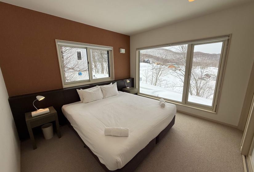 Spacious bedroom at Yutaka Townhouses featuring comfortable bed with crisp white linens, warm wood accents, large windows with Niseko snow views, and contemporary mountain lodge design.