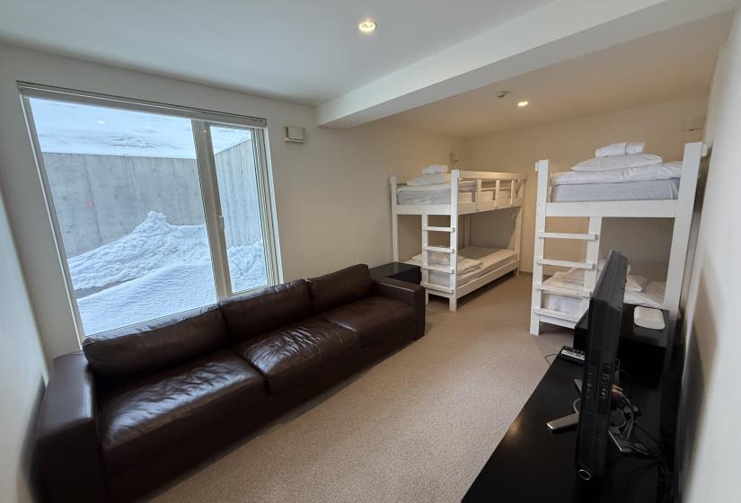Family bunk bedroom at Yutaka Townhouses featuring two white bunk beds, comfortable brown leather sofa, large window with snow views, and TV entertainment unit, perfect for children and teens during Niseko ski vacations.