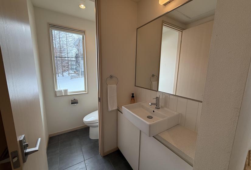 Bright bathroom at Yutaka Townhouses featuring white vanity with storage, wall-mounted mirror cabinet, modern toilet, window with natural light, and contemporary gray flooring, providing comfortable amenities for Niseko ski holiday guests.