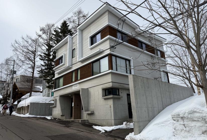 Exterior view of modern Yutaka Townhouses in winter featuring contemporary architecture with wooden accents, concrete elements, multiple levels, and balconies, surrounded by snow-covered landscape in central Niseko, just minutes from Hirafu ski lifts.