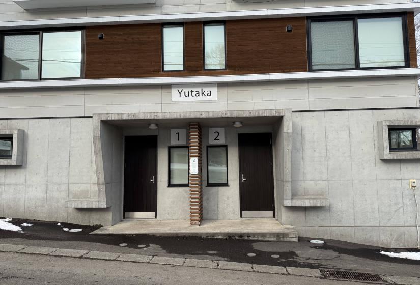 Front entrance of Yutaka Townhouses showing modern concrete and wood facade, twin entry doors with "Yutaka" sign above, and snowy street access, centrally located in Niseko for convenient ski holiday accommodation.