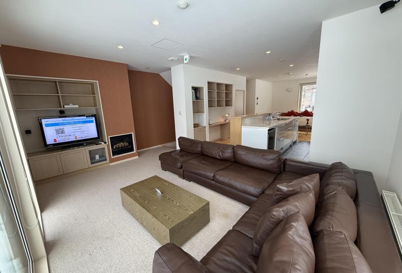 Spacious open-plan living room at Yutaka Townhouses with comfortable brown leather sectional sofa, wooden coffee table, flat-screen TV, warm wood accent wall, and bright contemporary design perfect for relaxing after skiing in Niseko.