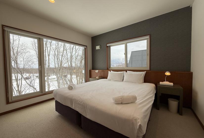 King bedroom at Yutaka Townhouses with luxurious bed, dark accent wall, large windows showcasing snowy forest views, bedside tables with reading lamps, and clean contemporary design for a comfortable Niseko ski holiday experience.