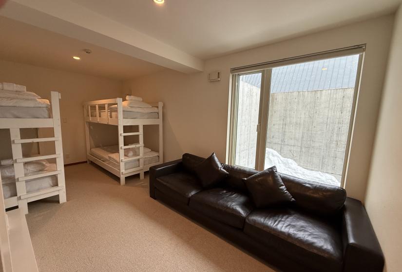 Family bunk room at Yutaka Townhouses featuring two white bunk beds, comfortable dark leather sofa, large floor-to-ceiling windows overlooking snowy Niseko landscape, and bright modern design perfect for children and teens during ski holidays.